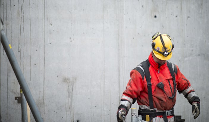 Byggnadsarbetare med gul hjälm och röda arbetskläder mot en grå betongvägg.