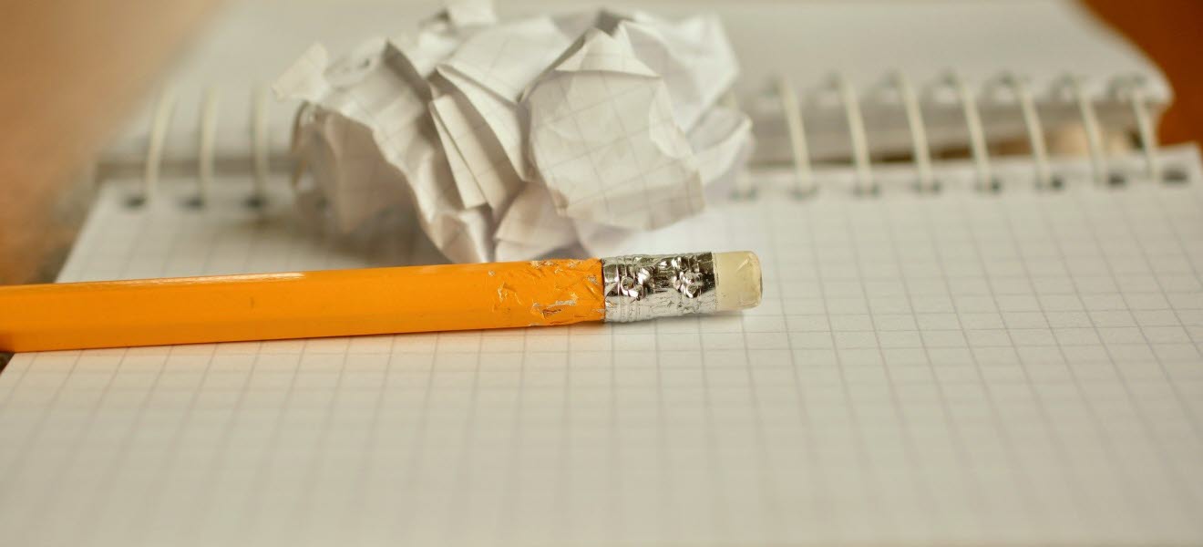 Orange penna med ett suddgummi på toppen som ligger på ett randigt kollegieblock. Bredvid ligger en ihopskrynklad pappersboll.